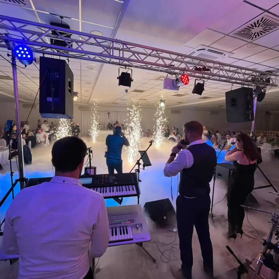 Bend nastupa u sali, dok mladenci plešu prvi ples na podiju, a fotograf ih fotografira.