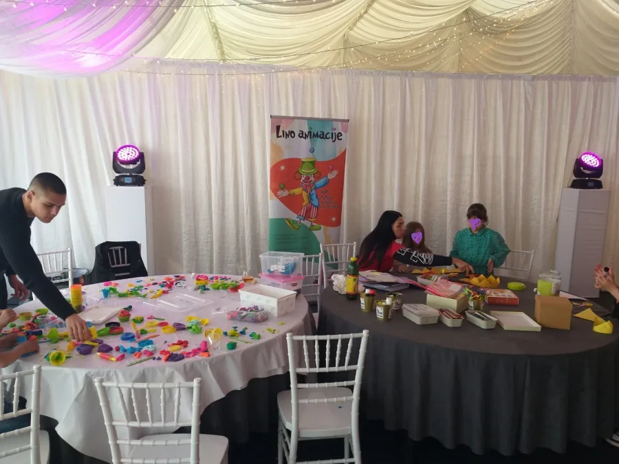 Djeca s animatorima u sali za vjenčanje stoje uz dva velika okrugla stola prepuna plastelinom.