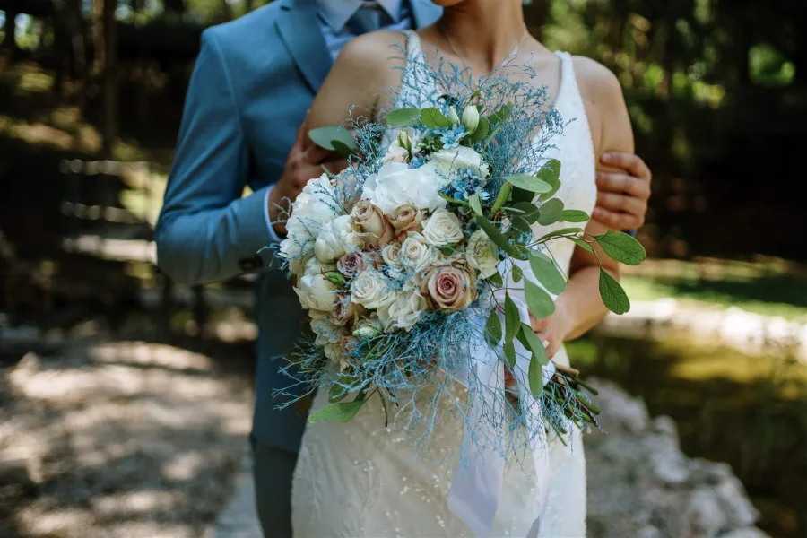 Mladenka stoji ispred mladoženje i drži buket u rukama, a on je drži za nadlaktice rukama.