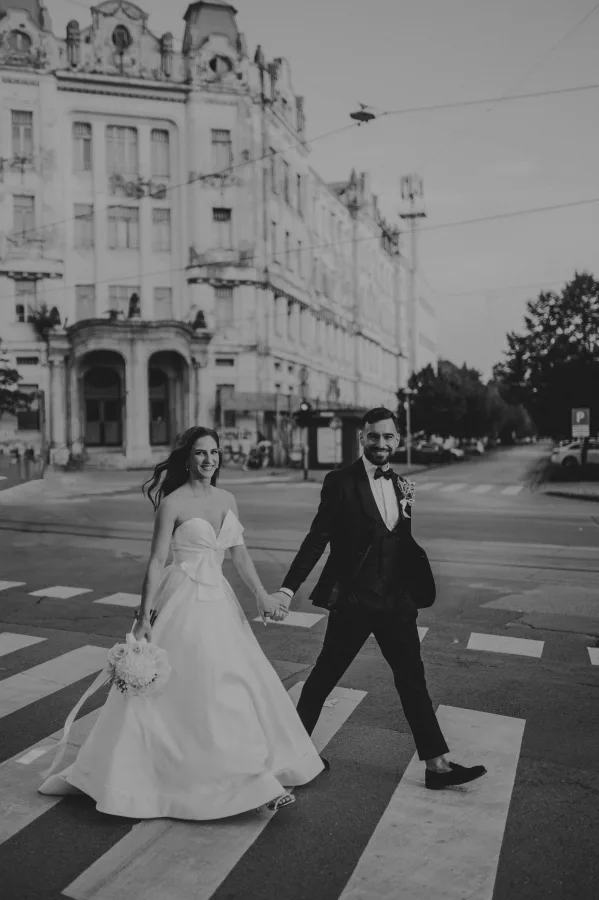 Mladenci prelaze pješački prijalaz držeći se za ruke i smiješeći se.