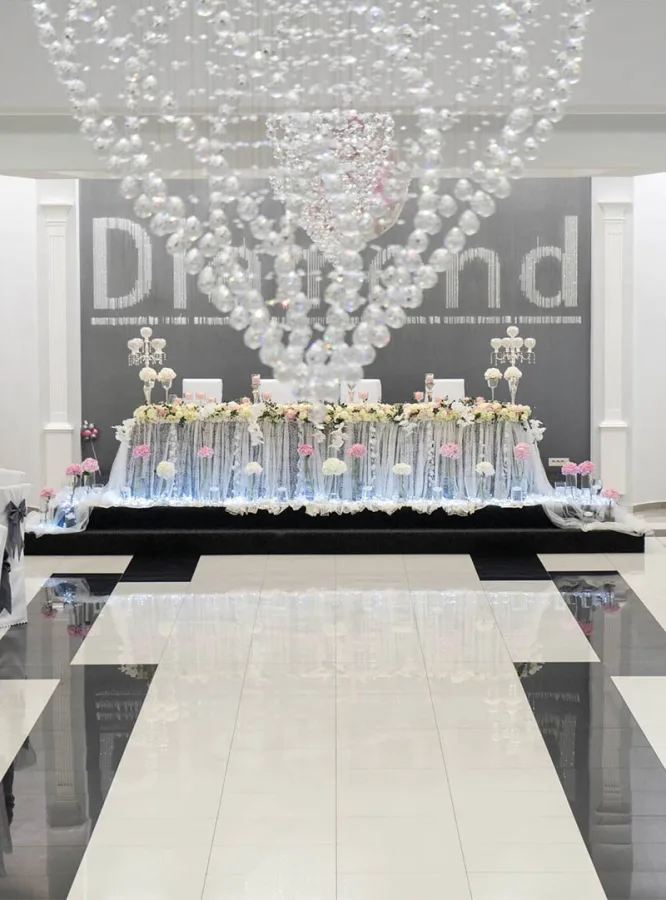 Kristalni luster usred podija sale za vjenčanje i stol mladenaca dekoriran cijećem.