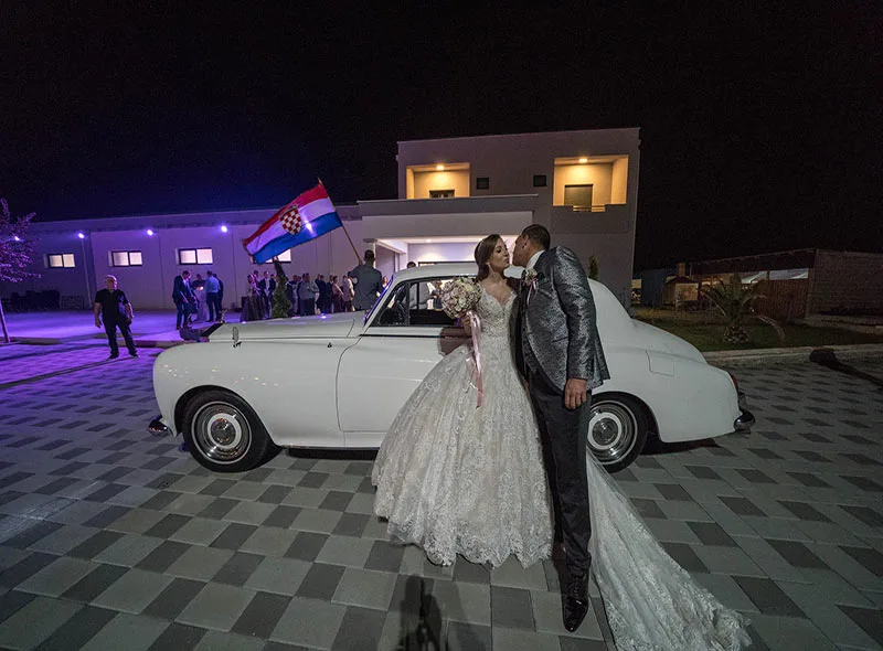 Mladenci skoro pred poljupcem, ispred sale za vjenčanje, naslonjeni na bijeli auto.