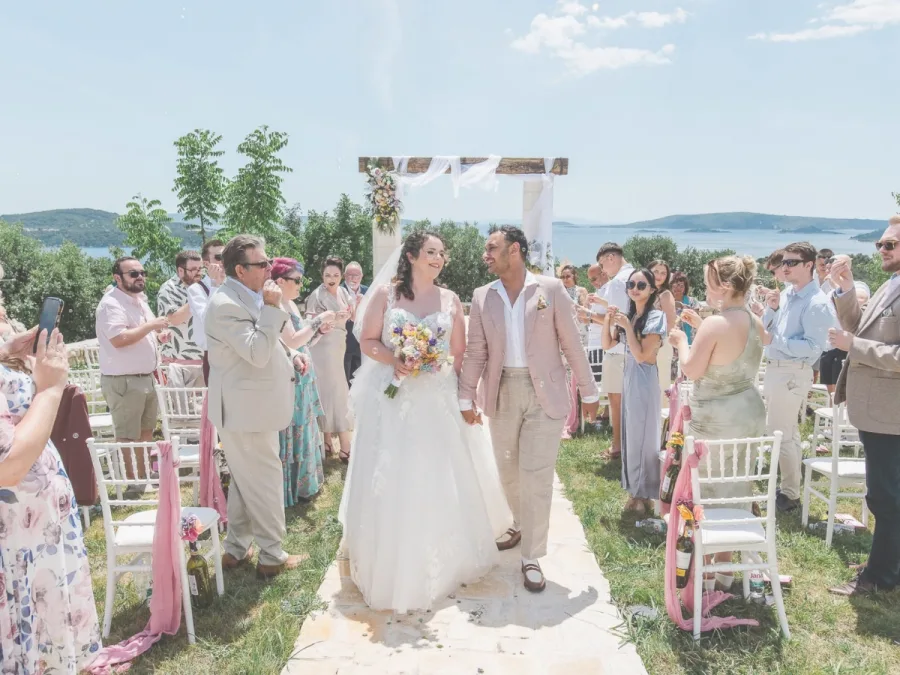 Mladenci hodaju stazom nakon obredne ceremonije u prirodu, okruženi obitelji i prijateljima, Mladenka drži šareni buket u ruci.
