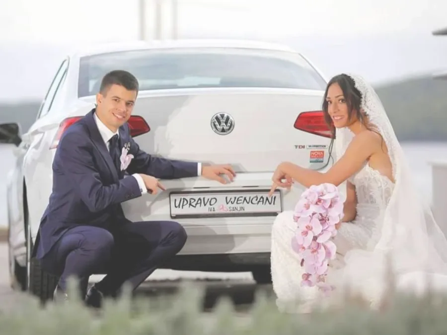 Mladenci čuče pokraj auta marke Volkswagen na kojem piše "upravo vjenčani". Mladenka u bijeloj vjenčanici drži ljubičasti buket u ruci.