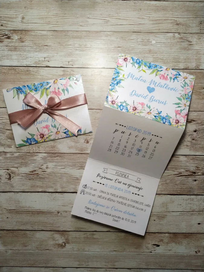 Pozivnica za vjenčanje s plavim i ružičastim cvijećem s imenima mladenaca u sredini i crvenkasto-ružičastom mašnom.