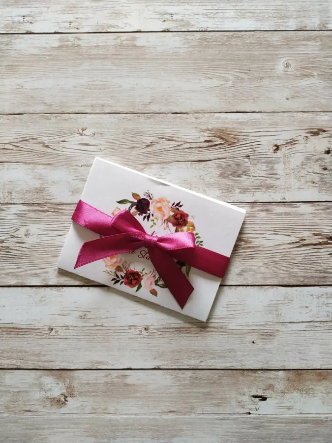 Pozivnica za vjenčanje s cvijećem i imenima mladenaca u sredini, a preko svega je ružičasto-crvena mašna.