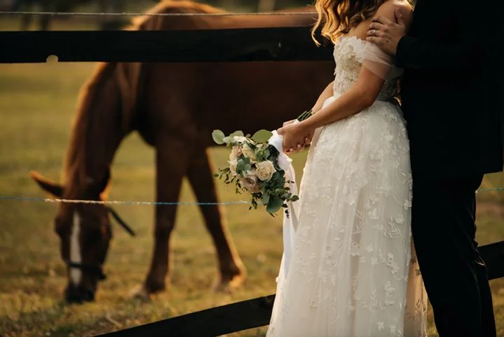 Mladenka u bijeloj vjenčanici drži buket dok konj pase travu u pozadini.