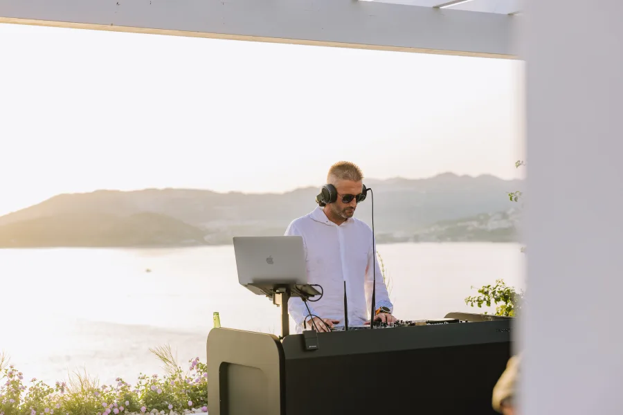 Dj sa slušalicama za miksetom dok je u pozadini predivan pogled na more uz zalazak sunca.