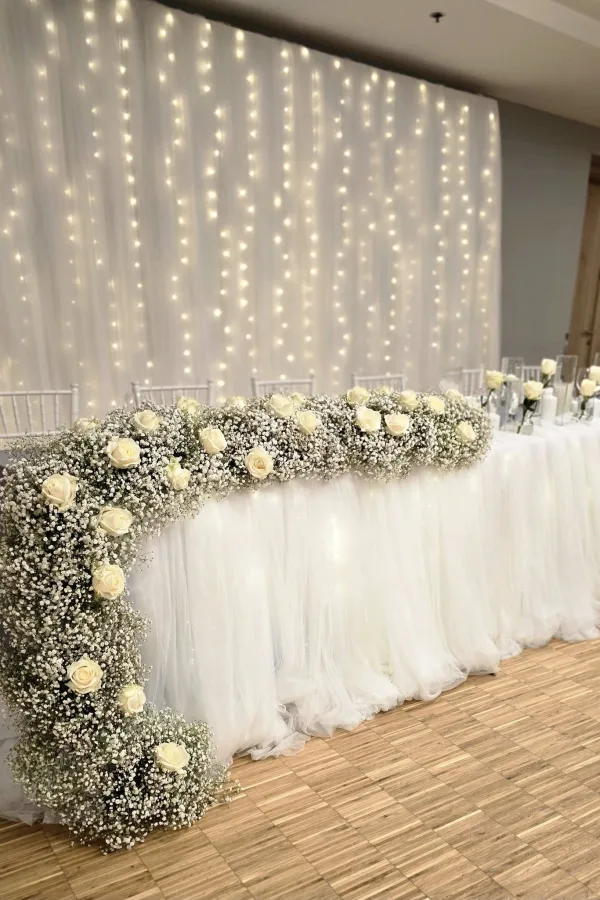 Bijeli stol za mladence u sali ukrašen bijelim cvijećem.