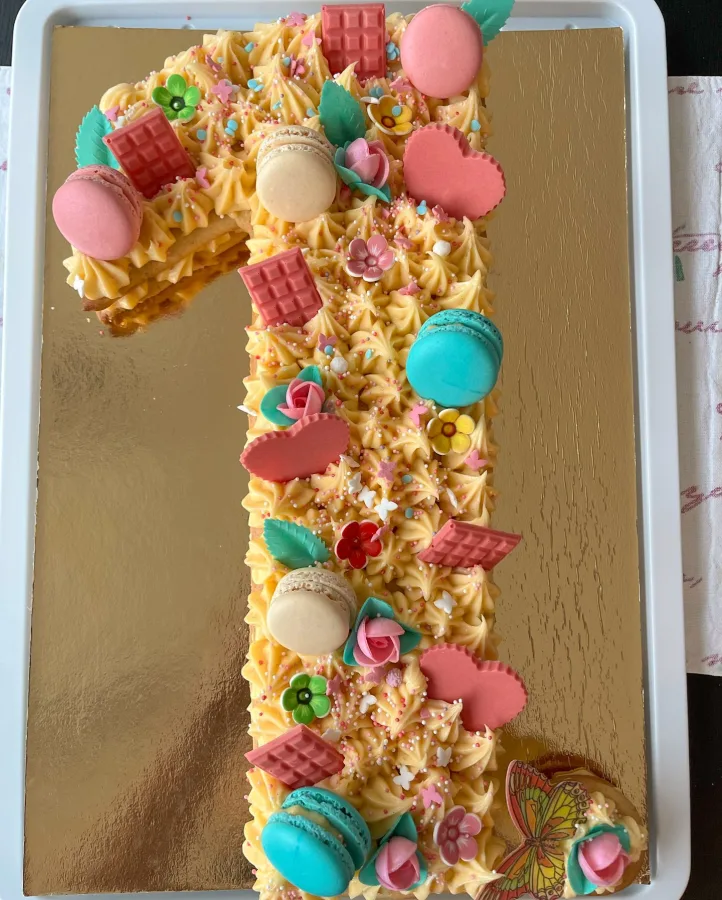 Torta u obliku brojke 1 s žutom kremom, rozim čokoladicama, makaronima i cvijetićima za prvi rođendan.