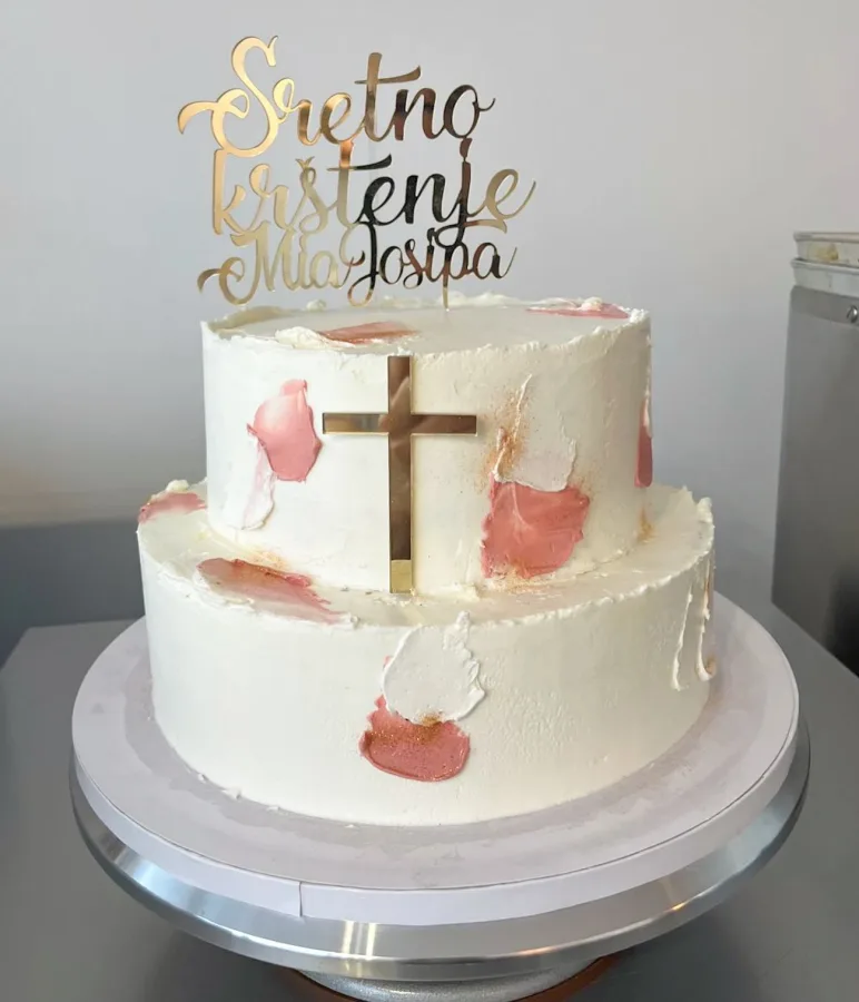 Bijela torta na dva kata s rozim detaljima, zlatnim križem i natpisom Sveto krštenje.