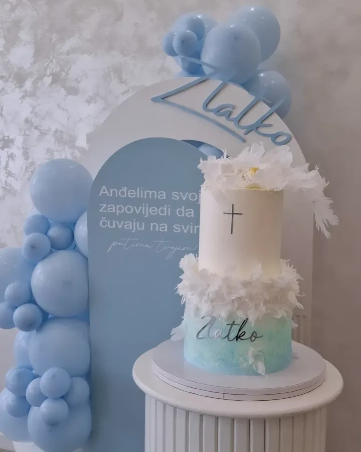 Torta za krštenje na dva kata s natpisom "Zlatko" na stalku pokraj dekoracije za krštenje. Prvi kat je plavi, drugi je bijeli i na oba kata je perje raspoređeno.