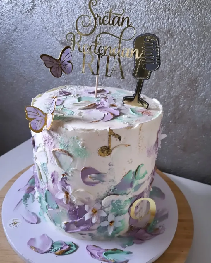 Bijela torta s ljubičastim i zelenim aticama od kreme, natpisom "Sretan rođendan Rita" , mikrofonom i eptirićima od papira.