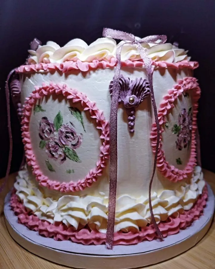 Bijela torta ukrašena ružičastom kremom i bijelom kremom te ljubičastim cvijećem i zelenim listovima.