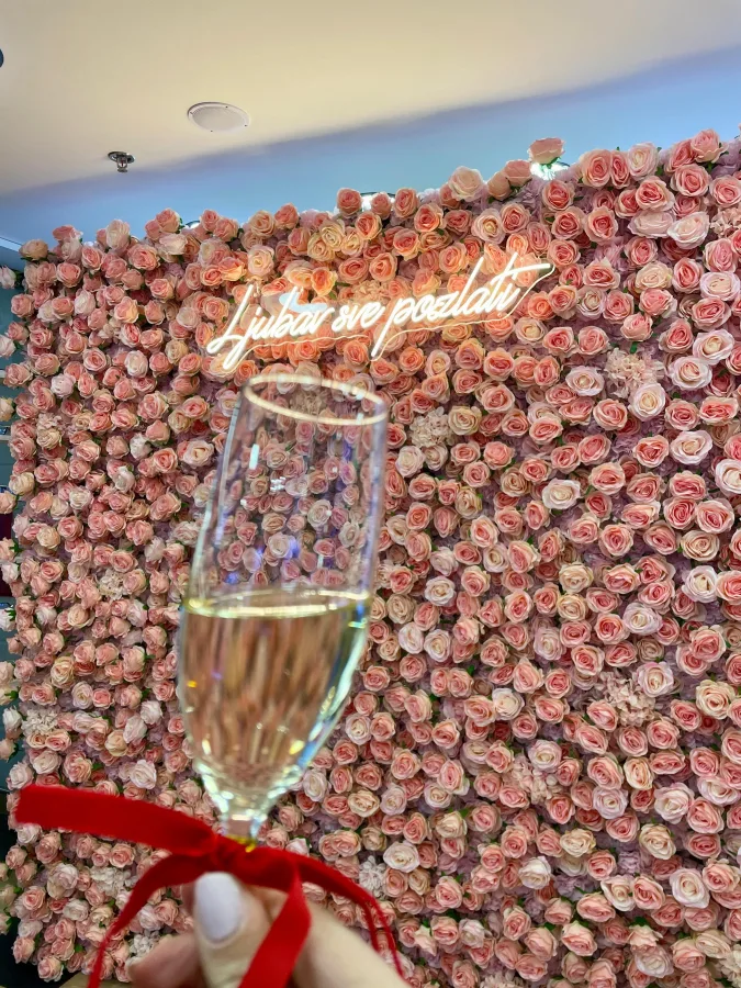 Zid s ružičastim ružama i neonskim natpisom 'Ljubav sve pozlati', idealan za fotografiranje na vjenčanju. Ispred zida ruka sa čašom šampanjca i crvenom mašnom.