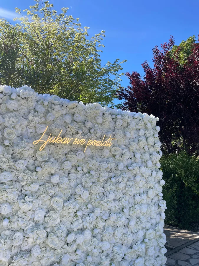 Zid s bijelim ružama i neonskim natpisom 'Ljubav sve pozlati', idealan za fotografiranje na vjenčanju.