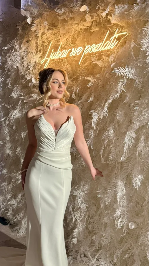 Mladenka ispred bijelog zida od perjem i neonskim natpisom 'ljubav sve pozlati', na vjenčanju.
